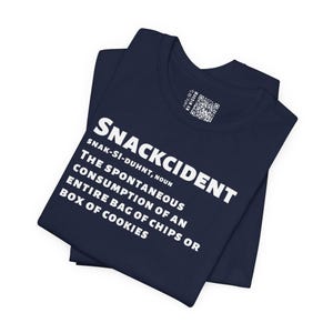 May include: A navy blue t-shirt featuring white text that reads SNACKCIDENT, SNAK-SI-DUHNT, NOUN, THE SPONTANEOUS CONSUMPTION OF AN ENTIRE BAG OF CHIPS OR BOX OF COOKIES. A small square QR code label is visible on the inside collar area of the garment.
