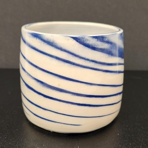 Pode incluir: Uma caneca cilíndrica de cerâmica com uma base branca lisa e uma série de riscas horizontais azuis finas, pintadas à mão, que envolvem o exterior. O interior da caneca é branco e o rebordo é rematado com uma linha azul fina.