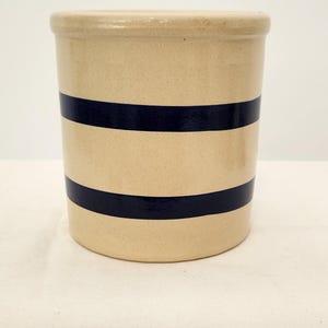 May include: A cylindrical ceramic crock with a smooth, light beige finish. The vessel features two horizontal, dark navy blue stripes encircling the body. It has a thick, rounded rim at the top and a flat base, maintaining a consistent, uniform shape.