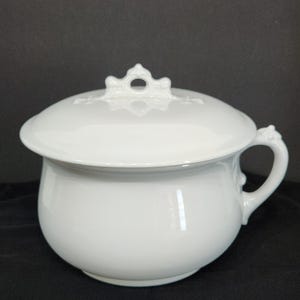May include: A white ceramic chamber pot with a lid. The pot has a rounded shape with a handle on the side and a decorative handle on the lid. The lid has a decorative design.