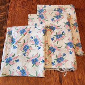 Puede incluir: Tres piezas de tela blanca con un estampado floral. El estampado presenta flores azules y rosas con hojas verdes. La tela está dispuesta sobre una superficie de madera.