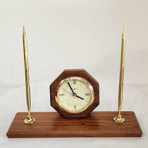 May include: A desk set featuring a rectangular wooden base with a dark brown grain. An octagonal wooden clock with a gold-tone bezel and cream face sits in the centre. Two gold-tone pens stand upright in holders on either side of the clock.