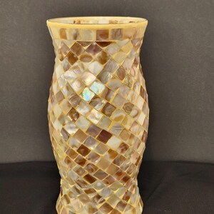 May include: A cylindrical vase featuring a mosaic pattern of small, square, iridescent shell tiles in shades of cream, beige, and brown. The surface has a glossy finish with a smooth, light-colored rim at the top and a matching base edge.