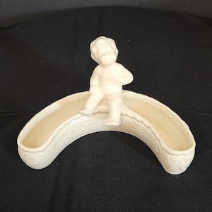 May include: Cream ceramic planter with a curved shape and two trough openings. A small, white cherub figure sits on top. The planter has decorative scrollwork along the edges. A decorative piece for the home.