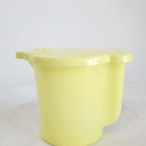 May include: A pale yellow plastic pitcher with a matching lid. The pitcher has a handle-like extension on one side and a spout-like feature on the other. The lid has a small tab for easy removal. The pitcher is designed for pouring liquids.