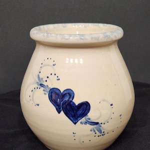 以下が含まれることがあります： 丸みを帯びたボディと水色の縁を持つ、クリーム色のセラミック花瓶。濃い青色のハート2つと青色の花のアクセントで装飾されています。家庭の装飾に最適な、魅力的で手作りの作品です。
