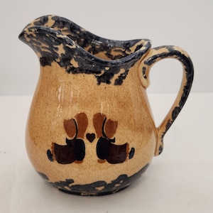 Pode incluir: Um jarro de cerâmica com esmalte bege salpicado e uma borda e base pintadas com esponja em azul escuro. A frente apresenta um design pintado à mão de dois cães castanhos frente a frente com um pequeno coração castanho escuro entre as cabeças.