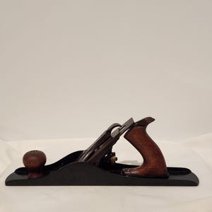 May include: A vintage hand plane with a black base, a dark wooden handle, and a wooden knob. The blade is made of metal. The tool is designed for woodworking and has a classic, well-worn appearance.