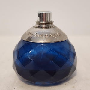 May include: A round, deep blue glass perfume bottle with a faceted, diamond-like texture. The bottle features a silver-toned metal collar engraved with the text Van Cleef & Arpels and a matching silver-toned spray nozzle at the top center.