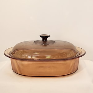May include: An oval-shaped amber glass casserole dish with a matching lid. The dish features vertical ribbed detailing along the exterior walls and a smooth, flat rim. The lid includes a central circular knob with a radial sunburst pattern.