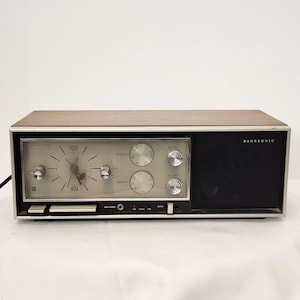Puede incluir: Un radio reloj AM/FM Panasonic rectangular con parte superior y laterales de madera. El frente presenta una esfera de reloj analógica plateada, dos diales de sintonización circulares, dos perillas de control y una rejilla de altavoz negra.