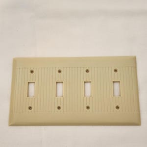 May include: A beige, four-gang light switch cover plate. The plate has four rectangular openings for the switches and four screw holes. The surface has a vertical ribbed texture.