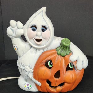 Peut inclure: Décoration d'Halloween en céramique représentant un fantôme souriant embrassant une citrouille. Le fantôme est blanc avec des yeux bleus et des joues roses. La citrouille est orange avec des accents verts et un visage sculpté. Un cordon d'alimentation sort de la base.