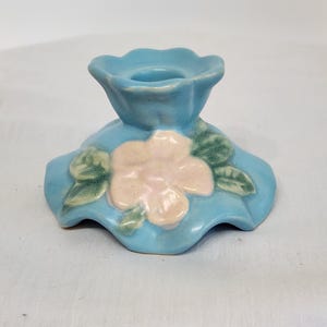 May include: A ceramic candle holder in a light blue colour, featuring a raised flower design in pink and green. The holder has a scalloped edge and a circular opening for a candle. A decorative piece for home decor.