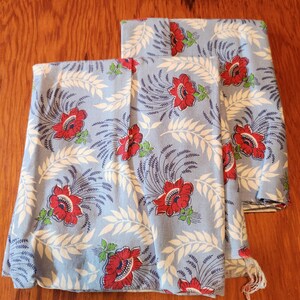 May include: Two folded light blue fabric pieces with a floral pattern. The pattern features red flowers, white leaves, and dark blue stems. The fabric is likely cotton or a similar material, suitable for sewing or crafting.