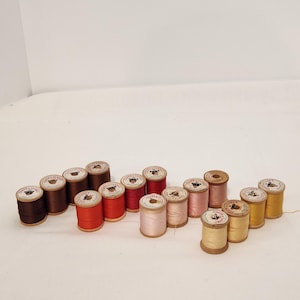 Könnte beinhalten: Eine Sammlung von Vintage-Garnrollen in verschiedenen Farben, darunter Braun, Rot, Rosa und Gelb. Die Spulen sind auf einer weißen Oberfläche angeordnet und zeigen ihre Holzsockel und Papieretiketten. Die Garnfarben variieren von dunkel bis hell.
