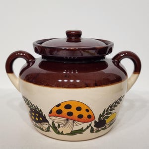 Puede incluir: Un azucarero de cerámica con tapa, en marrón y crema. El cuenco está adornado con un diseño de setas en naranja, negro y verde. Tiene dos asas y es adecuado para uso en la cocina.