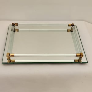 May include: A rectangular mirrored tray featuring a reflective glass base and a raised border constructed from clear glass rods. The rods are connected at each corner by metallic gold-toned hardware, creating a structured frame for the tray.