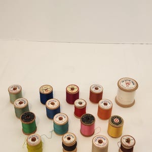 May include: A collection of vintage wooden spools of thread in various colors, including green, blue, red, and yellow. The spools are arranged on a white surface, showcasing the variety of thread colors and textures.