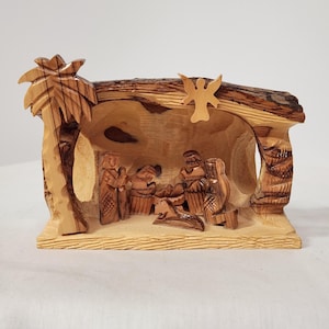 May include: Wooden nativity scene featuring carved figures of Mary, Joseph, and baby Jesus in a stable setting. The scene includes a palm tree and an angel, all crafted from light-colored wood. The figures are detailed and the overall design is rustic.