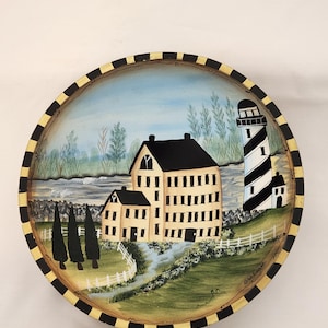 Op de afbeelding: Een rond decoratief houten dienblad met een handgeschilderd kusttafereel met een groot beige gebouw, een zwart-wit gestreepte vuurtoren, groene bomen en een wit hek. De rand heeft een zwart-geel geblokt patroon.