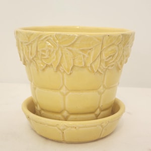 May include: A pale yellow ceramic flower pot with a matching saucer. The upper rim features a raised relief pattern of roses and leaves, while the body of the pot and the saucer display a grid-like texture of embossed squares with small diamond accents.