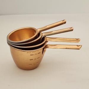 May include: Set of four vintage copper measuring cups stacked together. Each cup has a long handle and measurement markings. The cups are in varying sizes, with the largest cup labeled "ONE CUP TO TOP".
