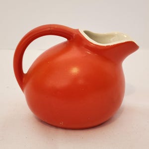 Könnte beinhalten: Ein runder Keramikkrug mit einer leuchtend orangefarbenen Außenseite und einer glatten, glänzenden Oberfläche. Das Gefäß verfügt über einen geschwungenen Griff an der Seite und einen breiten, offenen Ausguss mit einem kontrastierenden weißen Inneren.
