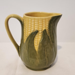 Peut inclure: Un pichet en céramique en forme d'épi de maïs avec une texture de grains jaunes sur la partie supérieure et des feuilles vertes enveloppant la base. Le pichet présente une anse incurvée sur le côté gauche et un bec verseur sur la droite.