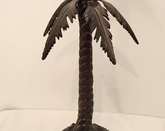 May include: A dark brown metal candle holder shaped like a palm tree. The trunk is textured with a repeating ring pattern, and the top features a circular candle cup surrounded by metal palm fronds with subtle green patina accents.