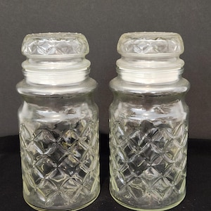 May include: Two clear glass canisters with matching lids, featuring a raised diamond-shaped lattice pattern on the body and a smaller diamond texture on the top of each lid. The containers have a cylindrical shape with a slightly flared rim.