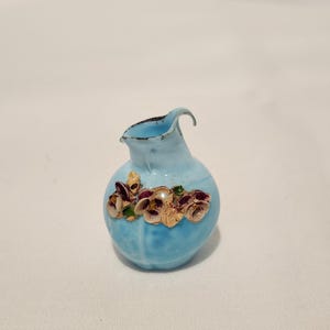 May include: A small, light blue ceramic pitcher with a curved spout and a round body. The pitcher is decorated with a cluster of raised, multi-colored floral designs. The background is a plain white surface.