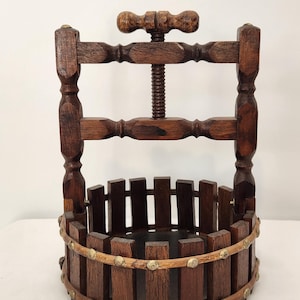 May include: A dark brown wooden fruit press featuring a cylindrical basket constructed from vertical slats held by a light-colored wooden hoop with brass studs. A central threaded screw mechanism with a top handle sits above the basket.