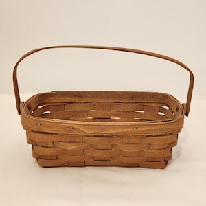 May include: A rectangular, woven brown basket with a curved handle. The basket is made of thin strips of wood, creating a textured surface. The handle is attached to the sides of the basket. The basket is empty and sits on a white surface.
