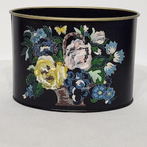 Könnte beinhalten: Schwarzer ovaler Mülleimer aus Metall mit goldfarbenem Rand. Der Mülleimer hat ein gemaltes Design mit einem Korb voller Blumen, Trauben und einem Schmetterling.
