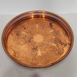 May include: A round copper-colored tray with an ornate, etched design. The tray has a decorative, pierced edge and a raised rim. The surface shows signs of age and wear, adding to its vintage appeal. A classic serving piece.