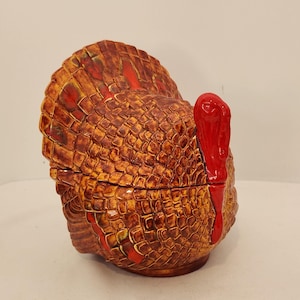 Peut inclure: Un récipient en céramique en forme de dinde avec couvercle, présentant un motif de plumes texturé en écailles dans des tons de brun, orange et jaune. La dinde possède une caroncule et un bec rouge vif brillant, avec une démarcation visible.