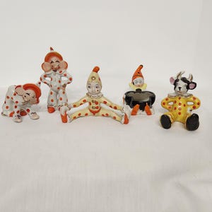 Puede incluir: Colección de figuras de cerámica vintage con diseños de payasos y animales. Las figuras incluyen un payaso en posición sentada, un payaso de pie, un payaso tumbado, una cabra y un payaso con una olla negra. Las figuras están pintadas con colores rojo, amarillo y blanco.