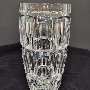 Puede incluir: Un jarrón de cristal tallado transparente con un diseño geométrico. El jarrón tiene una parte superior ancha y rectangular y una base acampanada. El cuerpo del jarrón está dividido en secciones con cortes verticales y arqueados, creando una apariencia texturizada. El jarrón mide unos 20 cm de alto.