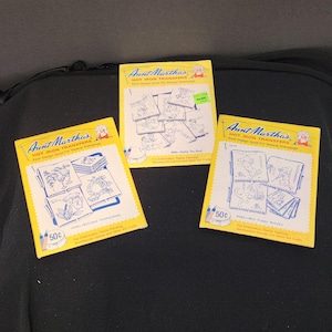 Puede incluir: Tres paquetes rectangulares de transferencias de planchado Aunt Martha's con bordes amarillos y centros blancos. Cada paquete presenta ilustraciones de líneas azules de varios motivos, incluyendo artículos de cocina, un pato y cachorros.