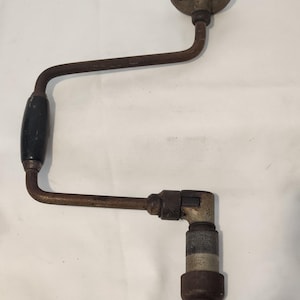 May include: A vintage hand brace drill featuring a rusted metal U-shaped frame, a black wooden rotating handle on the side, a black circular top knob, and a silver-toned metal chuck at the base for holding drill bits securely in place.
