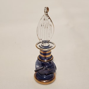 May include: A small, ornate perfume bottle with a clear, teardrop-shaped stopper. The bottle is navy blue with gold bands and trim. The stopper is clear and ribbed, adding a decorative element to the design. The bottle is likely made of glass.
