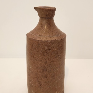 Può includere: Una brocca cilindrica in gres marrone con una superficie ruvida e maculata. Il recipiente presenta un collo stretto, un piccolo beccuccio per versare e un distinto rilievo orizzontale che circonda la parte superiore dove il collo incontra il corpo.