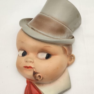 May include: A vintage wall hanging featuring a stylized baby face with a top hat and pipe. The baby has a red scarf and a white collar. The hat is gray and brown, and the face is painted with rosy cheeks and dark eyes.