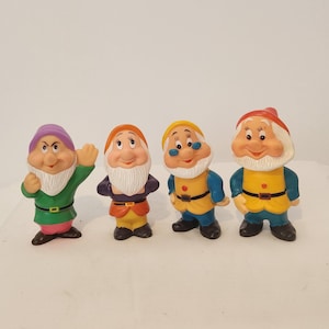 May include: A set of four dwarf figurines with white beards, each wearing a colorful outfit. The first wears a purple hat and green tunic, the second an orange hat and dark top, the third a yellow hat and blue shirt, and the fourth a red hat.