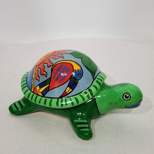 May include: A ceramic turtle ornament with a green body and a shell featuring a colourful bird and sun design. The turtle's shell is light blue with a green border. The figurine is a decorative item.