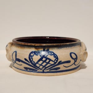 May include: A ceramic bowl with a cream-colored exterior and a dark brown interior. The bowl features blue floral and heart-shaped designs. Two small handles are on either side of the bowl.