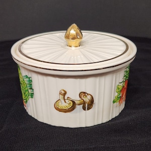 Puede incluir: Un cuenco blanco de cerámica con tapa y pomo dorado. El cuenco presenta un diseño acanalado y está decorado con ilustraciones coloridas de verduras y setas. Un ribete dorado acentúa la tapa y la base.