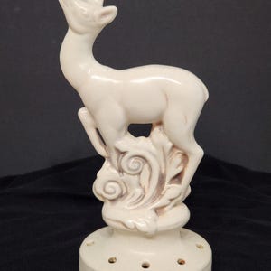May include: A cream-colored ceramic deer figurine with an upward gaze. The deer stands on a decorative base with floral accents and several small holes. The figurine is a decorative item.