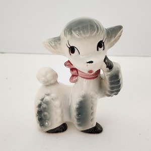 May include: A vintage ceramic lamb figurine. The lamb is primarily white with gray accents on its head, ears, and body. It has a pink bow around its neck and black hooves. The lamb has large, expressive eyes and a small, black nose.